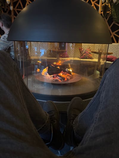 The image captures an intimate, low-angle view of a modern, glass-enclosed fireplace, likely in an indoor setting such as a lounge, restaurant, or home. The perspective is from someone sitting very close to the fireplace, looking down.
In the immediate foreground, two pairs of legs and feet are visible, belonging to individuals wearing dark trousers (possibly jeans) and dark, sturdy shoes or boots. The legs are positioned around the fireplace, suggesting they are seated and enjoying the warmth.
The central focus is the fireplace itself. It features a circular base where logs are actively burning, casting warm orange and yellow flames and glowing embers. The fire is contained within a clear glass cylinder, which allows an unobstructed view while maintaining safety. A dark, curved, semi-circular metal hood partially covers the top left section of the glass enclosure. Reflections are visible on the glass, showing distorted views of the surrounding room.
Through and around the fireplace's glass, elements of the interior decor can be seen. On the left side, beyond the fireplace structure, there's a partial view of a person's head and shoulders, suggesting another individual is also present. In the background, visible through the glass enclosure, there appear to be seating areas with cushions; one distinct cushion has a colorful, possibly striped or geometric pattern, and another reddish cushion is partially visible. Textured wall coverings, possibly quilted or patterned, can also be discerned in the background.
The wider scene includes decorative elements: a geometric wooden or metallic screen with a patterned design is visible in the top left and bottom left corners. Some decorative items resembling suspended paper lanterns or ornaments are also present near the top left. Green foliage, likely from a plant, adds a touch of nature to the top left corner.
The lighting is predominantly ambient, with the fire being a significant light source, casting a cozy glow. The overall atmosphere appears warm and inviting, indicative of an evening or nighttime setting. No visible text is present in the image.