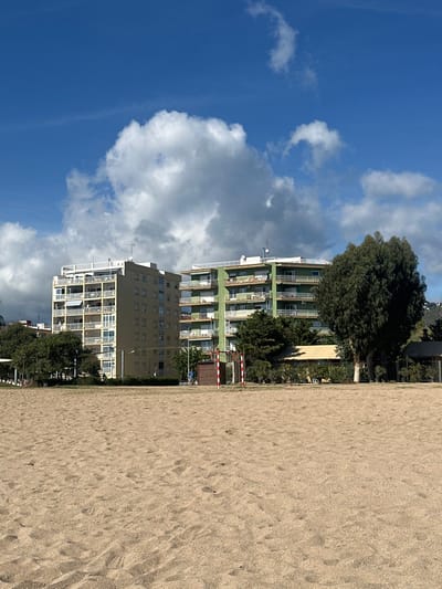 The image depicts a beach scene in Pineda de Mar, Spain, under a clear blue sky with scattered cumulus clouds. The foreground consists of an expanse of light brown sand with visible footprints. In the midground, a small, red and white striped goal post or barrier stands slightly left of center. Behind it, a narrow strip of green grass and low vegetation separates the sand from the urban background.

The background is dominated by two multi-story residential buildings. To the left, a light tan, rectangular building approximately eight to ten stories tall features numerous balconies, some equipped with white awnings, and a flat rooftop adorned with various antennas. To the right, a green, semi-circular building of approximately seven to eight stories also presents multiple balconies and a flat rooftop with antennas. A large, dense, dark green tree partially obscures the right side of the green building. Other smaller trees and bushes are visible between the sand and the buildings. Farther in the distance on the right, vegetated hills are visible.

A single individual, dressed in blue, is faintly visible in the midground, positioned to the left of the red and white goal, appearing to walk or stand. No other people or significant actions are discernible. The scene is illuminated by daylight, with shadows indicating sunlight from the left.