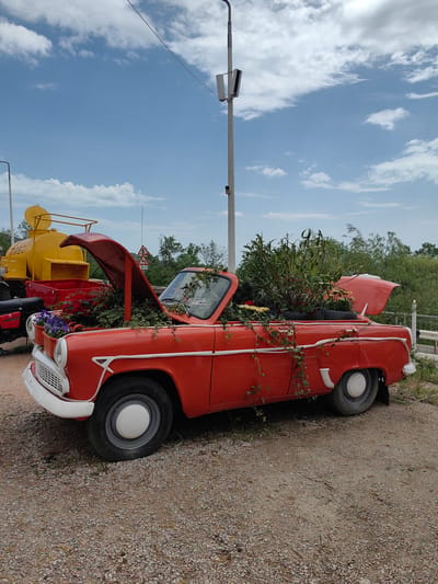 An orange Moskvitch car, repurposed as a flower bed, sits in Luchistoe, Ukraine. The hood and trunk are open, filled with various plants and flowers. The car is surrounded by a dirt and gravel ground. Behind the car, there's a yellow tanker and a white pole with lighting fixtures. A partly cloudy sky is visible above.