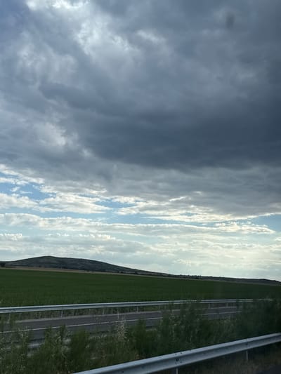 This is a horizontal, eye-level, medium shot of a rural landscape under a dramatic sky. The foreground features a highway with a silver guardrail running horizontally across the frame. Beyond the highway, a vast, green field stretches towards a rolling hill in the middle ground. The hill is covered in vegetation, and a small structure, possibly a tower, is visible on its crest. The background is dominated by a layered sky, with dark, heavy storm clouds occupying the upper portion of the frame, transitioning into lighter, wispy clouds and patches of blue sky towards the horizon. The lighting suggests it might be late afternoon or early evening. The scene depicts a natural environment with elements of human infrastructure. The location is identified as Polsko Padarevo, Bulgaria.