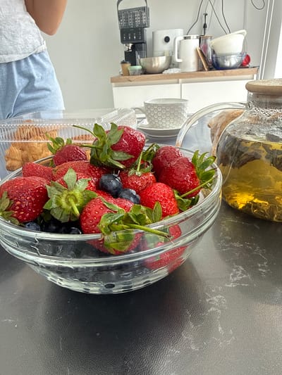 A close-up, rotated image shows a glass bowl filled with strawberries and blueberries. The bowl sits on a dark surface, possibly a countertop. Behind the bowl, a glass teapot filled with a yellow liquid (likely tea) is partially visible. Further to the left, a kitchen counter with various appliances and dishes is present. In the lower left corner, a person wearing light blue clothing is partially visible. The location is Olginskaya, Russia.