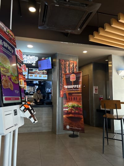 The image depicts the interior of a Burger King restaurant, possibly located in the Subang Jaya City Council, Malaysia. A self-ordering kiosk with a touchscreen display is prominently positioned in the foreground, on the left side of the frame. It shows images of burgers and other menu items. To the right of the kiosk, a tall banner featuring the Burger King logo and an image of a Whopper burger is displayed. Further back, a counter area is visible with a menu board above it and a cashier wearing a uniform and hijab serving a customer. Several tables and chairs are arranged in the dining area, and a high-efficiency air conditioner unit is installed on the ceiling. The restaurant interior is characterized by modern design elements, including grey tiled floors and walls, and a wooden slatted ceiling feature.
