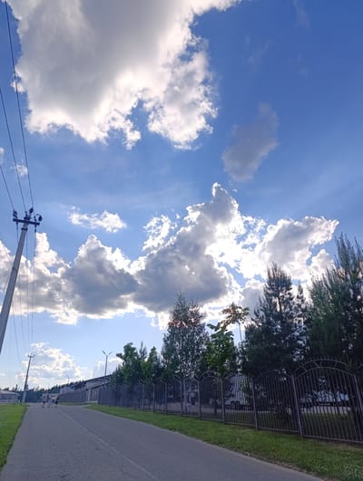 The image depicts an outdoor scene in Ivanovskoe, Russia, under a bright blue sky adorned with numerous large, fluffy white and grey cumulus clouds. The sun is partly obscured by these clouds, creating visible sunbeams and brightly illuminating their edges.
A paved road or pathway, possibly asphalt, stretches from the foreground into the distance, appearing to curve gently to the right. On the left side of the road, a concrete utility pole equipped with multiple power lines stands tall. Further down the road on the left, four small figures, likely people, are visible walking or standing.
To the right of the road, a dark, ornate metal fence runs parallel to the path, separating it from a row of green trees, including both coniferous (like pines) and deciduous varieties. Behind these trees and the fence, a long, low-rise building or complex is partially visible, along with several modern street lights. Scattered trees and another small structure can be seen in the distance on the left side of the road.
The scene conveys a calm, daytime atmosphere, likely late morning or afternoon, characterized by pleasant, partly cloudy weather. No visible text is discernable in the image.
