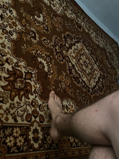 The image shows a person's bare leg and foot resting on a brown, patterned rug. The rug has intricate floral designs in shades of brown, beige, and white. The background is a light blue wall. There is a small red mark on the person's leg. The scene appears to be indoors, possibly in a residential setting in Soroca, Moldova.