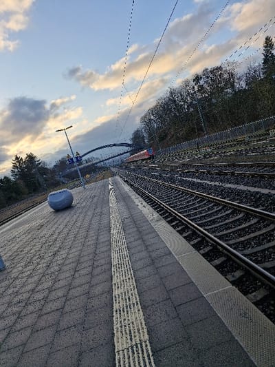 The image displays a train station platform in Niedernhausen, Germany, under a partially cloudy sky with hints of soft sunlight. The foreground features a grey, paved platform with a prominent tactile paving strip, composed of parallel raised lines, running along its length. To the left of this strip, a grey, rounded, roughly cylindrical object is positioned on the platform. To the right, multiple train tracks laid on ballast extend into the distance, running parallel to the platform. In the midground, a red and white passenger train car is partially visible on the tracks, positioned near an arched pedestrian bridge that spans over the railway. Overhead catenary lines crisscross the sky above the tracks. A tall, black lamppost and a blue sign indicating "5" and "6" are visible on the platform's left side, further into the scene. The background reveals a dense line of trees, predominantly deciduous and leafless, bordering a raised embankment alongside the tracks. No individuals are present in the scene, and no specific actions or interactions are depicted.