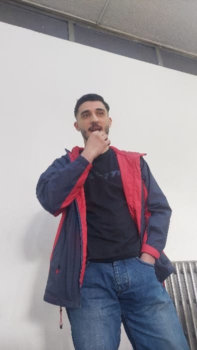 The scene depicts a male individual standing indoors within Al-Qaboun Municipality, Syria. He has short dark hair, a trimmed beard, and a mustache. His mouth is slightly open, and he is looking upwards and to the right. He is wearing a black t-shirt with a white graphic on the chest, over which he has an unzipped dark blue jacket with red interior lining visible along the collar and zipper. He is also wearing blue denim jeans. His right hand is raised to his face, with fingers touching his lower lip and chin area. His left hand is inserted into the left pocket of his jeans. The background consists primarily of a plain white wall. Above the white wall, a dark horizontal structure, possibly a window or partition, is visible, topped by a light grey, textured suspended ceiling with a visible grid. A vertically ribbed metallic object, potentially a radiator, is partially visible on the lower right side of the frame.