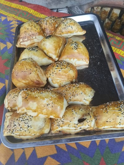 A close-up, eye-level shot shows a silver metal tray filled with golden-brown baked goods, likely samsa, arranged on a vibrantly colored rug with geometric patterns in red, yellow, blue, and green. The rug appears to be handmade. The samsa are topped with a mix of white and black sesame seeds and display varying degrees of browning and puffiness. Some samsa are triangular, while others are more rounded or rectangular. The tray rests on the rug, suggesting an outdoor market or food stall setting in Ala-Buka, Kyrgyzstan. In the background, out of focus, are additional baked goods stacked vertically, possibly in a tandoor oven, and a blurred glimpse of what might be more market stalls. There are no people visible in the frame.