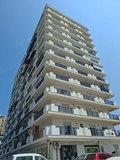 This is a low-angle, eye-level, medium shot of a modern, multi-story apartment building in La Manga Del Mar Menor, Spain, on a clear, bright, sunny day with a vivid blue sky. The building is tall and rectangular, with a white facade and numerous balconies. Each balcony is lined with dark blue railings, and many have retractable blue awnings extended. The architecture is characterized by repeated geometric shapes, with projecting elements on the balconies creating a layered effect. The lower floors appear to house commercial spaces, with storefront windows visible. One sign on the building reads "BLOQUE 2". There are air conditioning units mounted on the exterior walls. In the foreground, the top portion of a white van is visible on the left, and a red car is parked on the right. The overall impression is one of a densely populated coastal urban environment.