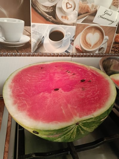 A half-cut watermelon sits on a stovetop. The watermelon is bright red with visible seeds and a green rind. In the background, a tiled backsplash displays a collage of coffee-themed images, including cups of coffee, croissants, and a "Good Morning" greeting. The scene is lit from above. The location in the city of Novoselytsia, Ukraine is not determinable from the image.