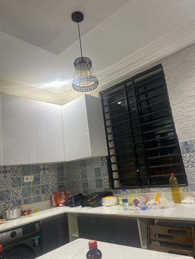 The image displays an interior scene, likely a kitchen or utility area, within a domestic setting in Adenta Municipality, Ghana. The photograph is oriented with a 90-degree clockwise rotation. A prominent hanging light fixture, featuring a decorative black metal cage design and an amber glass shade, brightly illuminates the space, suggesting artificial lighting is the primary light source, possibly indicating evening or nighttime.
The lower portion of a wall is adorned with an array of patterned tiles in shades of blue, gray, and white, exhibiting various geometric and abstract motifs. Above these tiles, the wall is plain white, and the ceiling features a textured surface. A large rectangular opening, possibly a window or a ventilation shaft, is secured with a black metal grille. Beneath this opening, a section of glossy white cabinets or storage units is visible.
A light-colored countertop occupies the right side of the frame, laden with several items. These include a tall yellow bottle of cooking oil, a clear plastic water bottle, and various plastic bags containing what appear to be groceries or household goods, such as a black trash bag and a clear bag with reddish contents. A red-capped bottle, possibly containing a beverage or sauce, is also present. Partially visible on the far right are a silver pot and part of a stove or cooker. A white electrical outlet with two sockets is installed on the tiled wall. Part of a wooden chair or stool with a cutout design is also visible on the right.
No people are present in the image, and no specific activity is explicitly taking place, though the arrangement of groceries suggests recent shopping or preparation for meals. There is no discernible text visible on any objects.