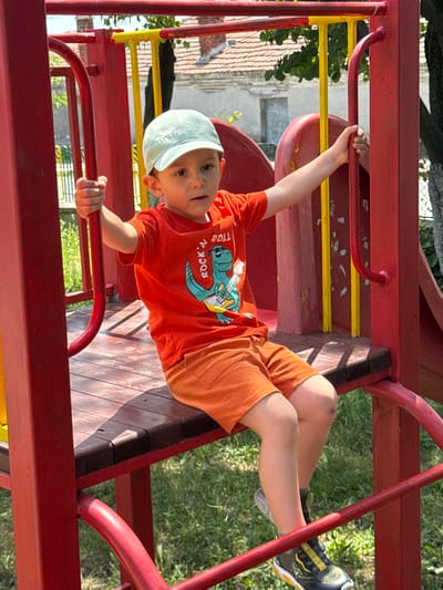 A young boy is sitting on a wooden platform of a red and yellow playground structure. He is wearing a light blue baseball cap, an orange t-shirt with a dinosaur graphic and the words "ROCK 'N ROLL" on it, and orange shorts. His legs are crossed, and he is holding onto a red metal bar with his right hand. The boy is looking off to his right. The playground structure is set against a backdrop of green grass, trees, and a building with a tiled roof. The image appears to have been taken on a sunny day. The location is Vinarsko, Bulgaria.