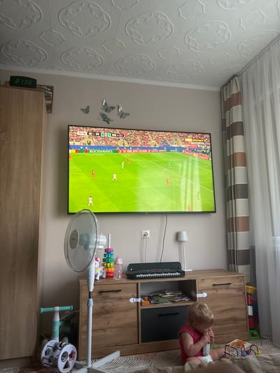 This media file captures an indoor scene, likely a living room in Krāslava, Latvia. The primary focus is a large television screen displaying a soccer match, indicated by the score "ClV 0 1 NOR" and the text "МАТЧ ПРЕМЬЕР" and "ПРЯМОЙ ЭФИР" which translates to "MATCH PREMIER" and "LIVE BROADCAST." The time displayed on a digital clock on a wooden cabinet reads "21:33," suggesting it is evening.
To the right of the television, a young child with blonde hair, wearing a pink shirt and a diaper, is seated on a beige surface, possibly a bed or sofa. The child appears engaged with a white object, possibly a toy. Next to the child is a wooden cabinet containing various items, including children's toys and bottles. A white oscillating fan is positioned in front of the cabinet, and other toys are visible on the cabinet and nearby shelves. Decorative butterfly decals adorn the wall to the left of the television. The room is illuminated by artificial light, and the presence of a digital clock and a television suggests a domestic setting.
