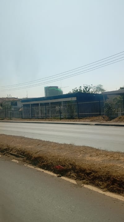 A street view captures a building behind a metal fence on a sunny day. The building appears to be a bus stop or small structure, with a taller, modern building with construction cranes in the background. A paved road runs in front, with dry grass along the side. Overhead wires stretch across the sky. The sky is light blue and clear, suggesting a warm day.