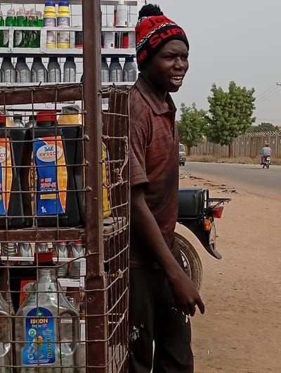 An African man in a red and black hat stands by a metal shelf loaded with motor oil bottles. He is wearing a striped shirt and black pants. The shelf is filled with various brands and types of oil. Behind him, a road is visible with a motorcycle driving on it. The background includes trees and a fence. The man appears to be a roadside vendor.