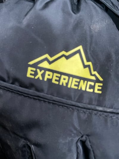 A close-up image displays a section of black synthetic fabric, consistent with material found on a backpack or outdoor gear, located in Pavlovo Podvor'ye, Russia. A yellow graphic is prominently featured, depicting a stylized mountain range above the capitalized word "EXPERIENCE." The fabric shows minor surface accumulation of light-colored dust particles, primarily noticeable in the upper right quadrant and along the bottom edge. A horizontal stitched seam is visible beneath the "EXPERIENCE" text, and the material exhibits slight creasing in various areas. No individuals or specific actions are depicted.