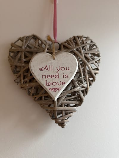 An indoor shot features a decorative heart-shaped object made of woven, natural-toned wicker, giving it a rustic appearance. This larger heart is positioned against a plain, light-colored background, likely a white or off-white wall.

A smaller, solid heart-shaped tag, colored white or off-white with a slightly distressed finish, is attached to the center of the wicker heart. The tag bears vertical text in a reddish-brown hue that reads, "All you need is love." A small, elaborate decorative motif, resembling a flourish or scrollwork, is printed beside the word "love." The tag is secured to the wicker heart with a piece of natural-colored twine or string.

To the left of the wicker heart, a segment of a slender, pink stick or dowel is visible, appearing to be connected to the heart, possibly as part of its hanging mechanism or a complementary decorative accent. The scene is illuminated by soft, even lighting, which emphasizes the textures of the woven materials and the painted wooden tag. There is no human activity shown; the image is a still life of this decorative item. The item is located in Hazlemere, United Kingdom.