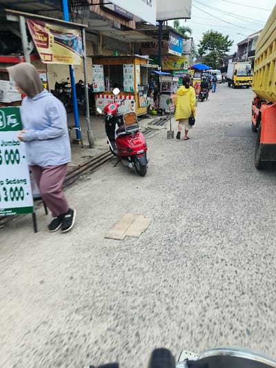 The image shows a street scene in Kecamatan Dayeuhkolot, Indonesia. A woman in a grey hoodie and pink pants is walking towards the camera while holding a sign with the words "Jumbo 5.000" and "Sedang 3.000". In the background, a woman in a yellow jacket walks away from the camera. A red scooter is parked on the right side of the street, and a large truck is parked on the far right side of the street. A sign with the word "Meval" is visible in the background. The street is paved with small gravel stones. There are buildings on the left side of the image and the sky is overcast. The street is relatively empty, with only a few people and vehicles present.