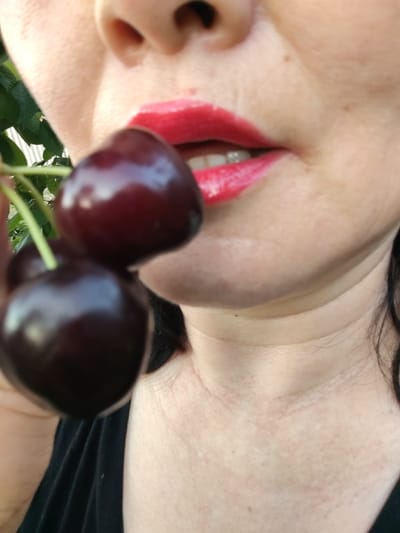This is a close-up, vertically oriented, medium shot of a person's mouth and chin area, holding a cluster of dark cherries. The person is wearing bright red lipstick and their teeth are visible as their mouth is slightly open. The cherries are dark red to almost black and appear ripe, with thin green stems connecting them. The skin tone of the person is light. The background is blurred, with some green foliage visible on the left side, suggesting an outdoor setting. The lighting appears to be natural, possibly from daylight, casting soft shadows. There is no visible text in the image. The focus is on the cherries and the person's lips, creating an intimate and suggestive feel. The overall impression is one of simple indulgence in fresh fruit.