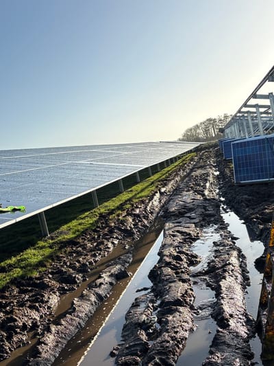 This image captures a ground-level view of a solar farm construction site in Grange Ville, Ireland, on a clear, sunny day. The foreground features a deeply rutted and muddy dirt track, filled with standing water reflecting the sky. This track runs diagonally from the lower left to the upper right. To the left of the track, a large array of installed solar panels, appearing slightly wet or reflective, extends into the distance, supported by metal posts above a strip of green grass. A small, bright green object is visible on the grass near the installed panels on the left. To the right of the muddy track, several blue solar panels are stacked vertically on the ground, adjacent to an unfinished metal framework structure awaiting panel installation. A partially visible yellow/orange object, possibly construction equipment, is present in the lower right corner, partially submerged in mud. In the background, a line of leafless trees borders the horizon under a bright, pale blue sky. No individuals are visible in the scene.