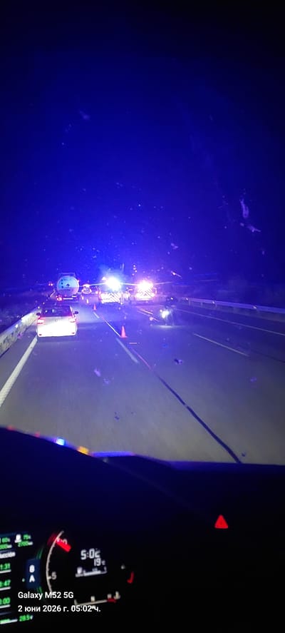 The image, rotated 90 degrees clockwise, captures a nighttime view from inside a vehicle looking forward onto a road. Bright white and blue lights intensely illuminate the left side of the road, indicating the presence of emergency services, maintenance vehicles, or construction activity. Several orange traffic cones are visible on the road surface, marking a work zone or an incident. The sky above is dark blue/black.

In the bottom right of the frame (when correctly oriented), a section of the vehicle's dashboard is visible. It displays digital information, including a clock showing "5:02" and a reading of "19", likely representing temperature. A red triangular icon, characteristic of a hazard warning light, is also visible.

Embedded in the image, text identifies the camera device as "Galaxy M52 5G". A timestamp reads "2 юни 2026 г. 05:02 ч.", which translates from Bulgarian to "June 2, 2026, 05:02 AM", confirming the early morning hour. The scene takes place in Pont de Molins, Spain, depicting an event or work in progress on the road during the predawn hours.