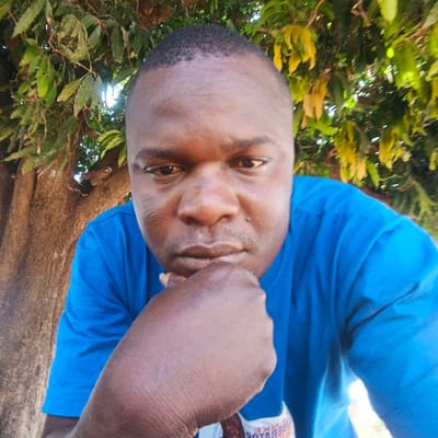 The image is a close-up portrait of a dark-skinned man, likely in his late 20s to 40s, captured outdoors. He has short dark hair and a serious or contemplative expression, with his right fist resting under his chin. He is wearing a plain blue t-shirt. In the background, a large tree trunk is visible on the left, and its branches with dense green and some yellowing leaves fill the upper and right parts of the frame, indicating a natural, outdoor setting in what appears to be a sunny daytime environment, possibly in Benin, Nigeria. Partial, mirrored white text is visible on his t-shirt near his right elbow, with letters appearing backward and difficult to fully decipher.