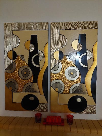 An indoor scene within the Municipal Unit of Acharnes, Greece, displays two identical rectangular abstract artworks mounted vertically side-by-side on a plain, light beige wall. Each artwork measures approximately 1 meter in height and 0.5 meters in width, depicting a geometric composition of stylized vessels or forms in shades of gold, black, silver, and tan. Portions of the artworks feature textured, possibly embossed or hammered metallic finishes, particularly within the gold and silver sections and the striped top and bottom borders. Below the artworks, a light brown, fluted or grooved ledge serves as a decorative surface. On this ledge, two red glass votive candle holders, each with dark intricate patterns, are positioned symmetrically at the left and right edges. Centered between them is a red rectangular tray containing four small, unlit red pillar candles arranged linearly. No individuals are present.