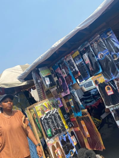 The image depicts an outdoor market stall in Aiyetoto-Asogun, Nigeria, under a clear, bright blue sky. A light-colored fabric canopy extends diagonally across the top right, providing shade for the stall. The stall primarily displays numerous packaged hair extensions and braids of various colors and styles, hanging and stacked. Visible brands include "X-Pression," "Darling," "Rich Braid," "Superstar," "Hairkey," and "Nectar." Hair colors range from black and dark brown to auburn and mixed tones.

A female individual is positioned in the lower left foreground, wearing a rust-colored short-sleeved top and a dark baseball cap. She stands facing slightly right, with her gaze directed off-camera to the right. Her left arm is visible, resting near the merchandise. In the background, another light-colored tent or large umbrella is partially visible to the left of the main canopy, and stacks of folded textiles, likely clothing, are discernible beneath the market stall structure. A portion of a white vehicle is visible further back in the mid-ground. The scene is illuminated by strong daylight.