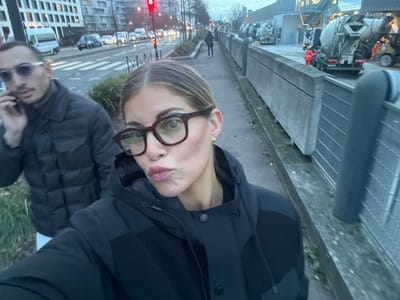 The image captures a selfie featuring two individuals outdoors, likely in an urban or semi-urban environment. The perspective is from slightly above, looking down, and the image is originally rotated 90 degrees counter-clockwise.

In the foreground, slightly to the left, is a person with fair skin and light-colored hair, pulled back, wearing brown-rimmed glasses. This person is looking directly at the camera with puckered lips, a common expression for a selfie. They are wearing a dark, possibly black, quilted or puffy jacket over a darker top. A silver earring is visible in their left ear.

To the right of this person, partially visible, is another individual. This person is also wearing a dark, possibly black, quilted or puffy jacket and dark sunglasses. Their head is tilted slightly, and they are also looking towards the camera. Their features are less discernible due to the sunglasses and partial cropping.

The background is split, showing different elements of the environment. On the left side of the frame (when correctly oriented), there appears to be a construction site or an industrial area. Heavy machinery, possibly including a cement mixer or similar large equipment, is visible. There are concrete structures, possibly barriers or foundations, and metal grates or fencing. The ground is paved, suggesting a sidewalk or service road.

On the right side of the frame, behind the individuals, the scene transitions to a typical city street. A road with white markings is visible, along with a red traffic light that is illuminated. Cars are parked or moving on the street. In the distance, there are several multi-story buildings, likely residential or commercial structures, lining the street. The sky above is overcast, and the overall lighting suggests either late afternoon, early evening, or a heavily clouded daytime. No discernible text is visible in the image.