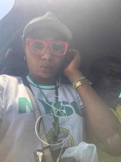 The image shows two individuals inside what appears to be a vehicle. In the foreground, a female with dark skin is prominently featured, looking directly forward. She wears a light green baseball cap, large red-framed clear glasses, and a white t-shirt with the green "NYSC" (National Youth Service Corps) logo and emblem. White earphone wires are draped over her neck and chest. On her left wrist, she wears a gold-colored wristwatch and a beaded bracelet. Her right hand is raised near her ear. To her right and slightly behind, a male with dark skin is partially visible, wearing a yellow shirt, with his head turned slightly away. Bright sunlight from the upper left creates significant lens flare and strong highlights across the scene, particularly on the female's cap and the upper left of the frame. The interior environment is dark, consistent with a vehicle's cabin. The specific city of Bundot, Nigeria, is not identifiable from any visual cues within the image.