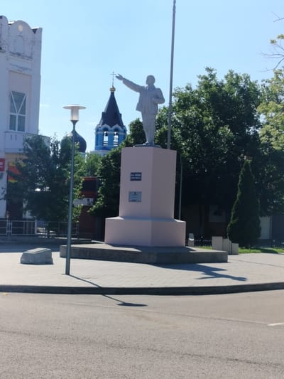 In Labinsk, Russia, a statue of Vladimir Lenin stands prominently in a public square, facing left with his right arm extended. The statue is made of a light-colored material and is mounted on a tall, light-pink pedestal. A plaque is visible on the pedestal. The statue casts a distinct shadow on the paved ground.
The square features a paved walkway and a street. In the background, a white building with arched windows is situated to the left. A bright blue church bell tower with a dark dome and a golden cross rises behind the statue, partially obscured by trees. Dense green trees surround the statue and the buildings, suggesting a summer day with clear, blue skies. A modern streetlamp is positioned to the left of the statue. To the right of the statue, a dark green, conical evergreen tree stands tall. The overall scene depicts a public space in Labinsk, likely a central area featuring historical monuments and civic buildings.