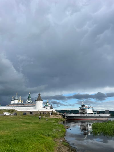 In Makar'evo, Russia, a cargo ferry, labeled "СП-35," navigates a body of water. The ferry, a gray and white vessel with red accents, is positioned near the shore, where tall green reeds grow. On the opposite bank, a historic complex featuring white walls and multiple domed churches with golden crosses stands against a dramatic, cloudy sky. Several cars are parked along the road next to the complex, and a few individuals are visible on the grounds. The water reflects the sky and the ferry, creating a mirrored image.