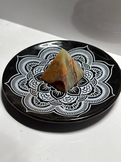 A small, polished, multi-colored gemstone pyramid, exhibiting bands of green, white, and reddish-brown, rests on a black, circular dish adorned with a white, intricate mandala-like design. The dish is positioned on a white surface, with a subtle shadow cast by the object and dish. The background is a plain, white, wall-like structure that angles upwards. The image is brightly lit, with a slight glare visible on the dish and the white surface. The scene depicts a still life arrangement of decorative objects. There is no discernible location in Gurkovo, Bulgaria.