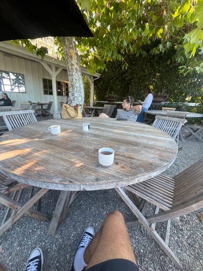 The scene depicts an outdoor dining area in Summerland, United States, characterized by a casual, rustic ambiance. In the foreground, a weathered, round wooden table occupies the majority of the frame, with the observer's legs and feet visible beneath it; the observer wears black shorts, white socks, and black Converse sneakers. On the table, two white ceramic mugs containing dark liquid are present, along with a small salt and pepper shaker set. Surrounding the table are several wooden slatted chairs.
The ground consists of light-colored gravel. Overhead, a large black patio umbrella is partially visible, and abundant green foliage from a large tree, likely a sycamore given its mottled bark, provides shade, casting dappled sunlight on the table.
In the mid-ground, a white-painted building with multiple windows, featuring horizontal paneling, stands to the left. A woman with dark hair, wearing a dark top, is seated at a table next to this building. To the right of the large tree, another man is seated at a wooden table, wearing a horizontally striped shirt and sunglasses resting on his head; he is looking towards the left. Behind him, a woman wearing a white top is standing, seemingly interacting with a mobile device. Additional wooden tables and chairs are scattered throughout the background, amidst dense green hedges and decorative planters. The lighting suggests a bright, sunny day.