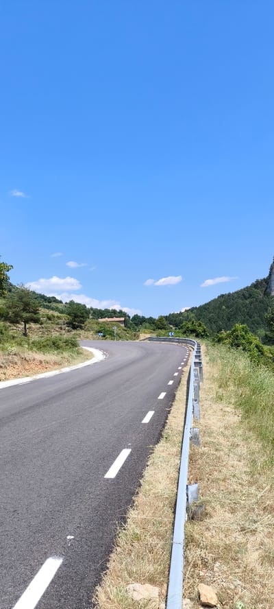 The image shows a curved asphalt road with white dashed lines on the right side. A silver guardrail runs along the right edge of the road, separating it from dry, yellowed grass and rocks. The road is flanked by a grassy, sloped hillside on the left, with scattered trees and bushes. In the background, there is a building with a red-tiled roof visible among a cluster of trees on a higher part of the hill. Further in the distance, a dense forest covers a mountainous landscape under a clear blue sky with a few scattered white clouds. There are no people or vehicles visible in the scene. This is a scene from the outskirts of Berga, Spain, a town located in the Berguedà region of Catalonia.