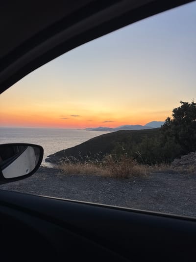 A scenic view of the coast is captured from inside a vehicle, likely in Glavatičići, Montenegro, during sunset. The sky exhibits a gradient of warm oranges and yellows near the horizon, transitioning to a soft blue overhead. The sea stretches out to meet a distant, hazy mountain range. In the foreground, dry grass and shrubs dot the rocky terrain alongside the road. A car's side mirror is visible on the left, reflecting a portion of the scene. The interior of the car, including the door frame, is also visible, framing the landscape. The overall impression is one of a peaceful, picturesque coastal drive at dusk.