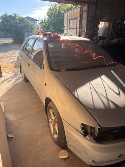 The image depicts a light-colored hatchback car, likely a Nissan Almera, parked inside a garage or workshop. The car appears to be undergoing bodywork or painting preparation, evidenced by a thin layer of primer or primer dust covering its exterior. A bright red, coiled air hose is draped over the roof and windshield of the car, with its ends connected to what appear to be pneumatic tools on the roof.

The garage interior is dimly lit, with sunlight streaming in from the left, casting long shadows across the car and the concrete floor. The walls are made of brick and concrete. Tools and equipment are visible in the background, suggesting an active workspace. Outside the garage, on the left side of the image, a dirt driveway leads to a residential area with a house and trees. The presence of what looks like sandpaper discs on the floor further indicates ongoing automotive work. The scene is set in Taman, Russia. No individuals are visible in the image.