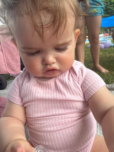 This is a close-up, vertically oriented, eye-level shot of a baby girl lying down outside. She is wearing a pink, ribbed onesie and her eyes are closed, giving her a peaceful expression. Her light brown hair is messy and falls across her forehead and cheek. In the background, out of focus, are the legs of an adult wearing blue shorts with a fish pattern, suggesting a casual outdoor setting. Further in the background, a blue umbrella and a portion of a pool with a purple inflatable toy can be seen. The presence of grass indicates an outdoor environment. The lighting suggests it is daytime, likely a sunny day, suitable for outdoor activities. No visible text is present in the image. The setting appears to be a backyard or park area, possibly near a swimming pool, and the activity is a baby resting outdoors.