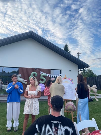 A group of people are gathered in a grassy backyard in Simagino, Russia, in front of a modern white house with a dark roof. The occasion appears to be a birthday celebration, indicated by large number balloons spelling "35" and a banner that reads "ДЕНЬ РОЖДЕНИЯ" (Birthday in Russian). Several individuals are interacting, with some conversing and others looking at a crossword puzzle displayed on an easel. In the foreground, a person wearing a black t-shirt with white lettering is seated. The sky above is partially cloudy, with scattered white clouds against a blue background. The environment is a well-maintained outdoor space, suggesting a residential property.