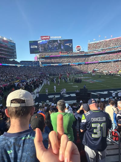 Levi's Stadium packed with spectators, a football field in view, and a large screen displaying the action. A Pepsi sign is visible atop the stadium. Fans are seated, some wearing team jerseys. The picture is taken from within the crowd, showing part of a person's head, hand, and the back of someone in a Seattle Seahawks jersey with Chancellor and the number 31 on it. The photo appears to be taken during a football game.