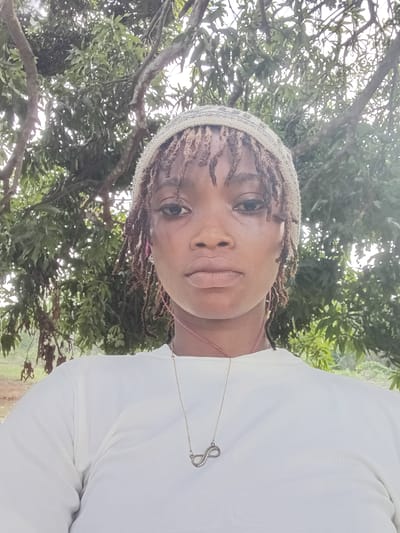 A close-up, slightly rotated portrait features a person, likely a woman, positioned to the right of the frame, looking directly at the camera. She has dreadlocks, partially covered by a light-colored, possibly knitted or crocheted beanie or headwrap. Her expression is neutral to slightly serene, with full lips and visible dark eyes. She is wearing a light-colored, long-sleeved top and a silver-colored necklace with an infinity symbol pendant, which appears to be embellished with small, sparkling stones.
The setting is outdoors during the daytime, with ample natural light filtering through the background. The background consists of dense, lush green foliage and tree branches, strongly indicating a natural environment such as a garden or a treed area. Small green fruits, consistent with mangoes, are discernible among the leaves, suggesting a tropical climate in line with the provided location context of Ogbe, Nigeria. No visible text is present in the image.