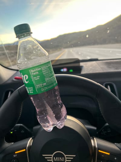 The image depicts the interior of a vehicle, specifically a Mini, with a clear plastic Sprite bottle resting vertically on the black leather steering wheel. The bottle is partially filled with a purple liquid and shows condensation on its exterior. Its green label displays "Sprite," "Nutrition Facts," "Calories 200," and "CA CRV." The steering wheel features a "MINI" logo, a "SET" button, and an illuminated orange indicator light. A heads-up display unit with green indicators is visible on the dashboard beyond the steering wheel. Through the vehicle's windshield, a multi-lane road or highway extends into the distance, flanked by arid, mountainous terrain consistent with the region around Reno, Nevada. The sky is bright, with sunlight causing glare on the windshield and horizon. No individuals are visible.