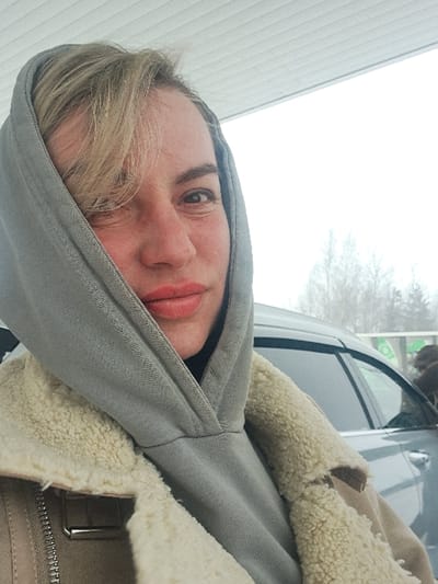 A person wearing a gray hooded sweatshirt and a tan shearling-lined jacket is positioned near a car with a light-colored building visible in the background. The individual's face, slightly red, is prominent within the hood. A tree with bare branches is seen through the car window. The photograph appears to be taken during the day with overcast lighting.