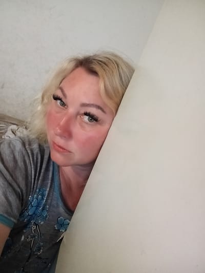 A blonde woman with heavy eyelashes leans against a light-colored surface, with her face partially obscured. She is wearing a grey t-shirt with a blue floral pattern and appears to have flushed cheeks. The background consists of a plain wall and a hint of fabric or bedding. The image is taken indoors. The image was taken in Kostiantynivka, Ukraine.