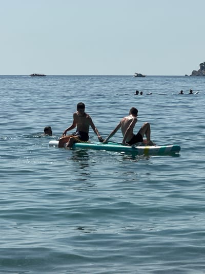In the waters near Đurovići, Montenegro, two young males are engaged in water activities. One is sitting on a paddleboard, his legs bent and arms resting on his knees. The other, partially submerged, is swimming nearby. In the background, several other individuals are also in the water, some with paddleboards. Further out, two boats are visible on the horizon, under a clear sky. The sea is calm with gentle ripples.