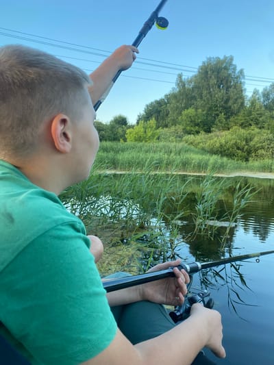 A boy, seen from the back and right side, is holding two fishing rods by the water. He is wearing a green t-shirt and dark pants. The boy is in the foreground and his face is turned to the right, looking towards the water. The water is calm and reflects the sky and trees. There are reeds growing in the water. In the background, there are trees and a clear blue sky. The scene appears to be set in a natural environment, possibly a lake or river, and given the context of the city of Afanasyevo, Russia, it is a rural setting outside of the city. The lighting suggests it is either early morning or late afternoon. There is no indication of any other people or animals in the image.
