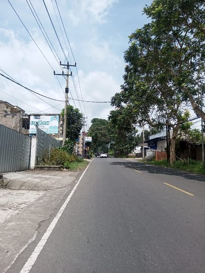 The image displays a straight asphalt road in the city of Khajuripada, India, under a partly cloudy sky. The road features a solid white line on its left edge, delineating it from a cracked concrete sidewalk, and broken yellow lines indicating two traffic lanes further in the distance.
On the left side, a corrugated metal fence runs parallel to the road. Behind this fence, a two-story building with a light-colored facade is visible. A large billboard is mounted on this building, featuring the prominent Indonesian text "ANANTA UMROH" and "23 Jutaan," alongside an image of a man in a white shirt. A wooden utility pole, heavily strung with multiple overhead power and communication lines, stands centrally on the left. Scattered green foliage and small trees are present along this side.
On the right side of the road, a dense line of large trees with green leaves, some bearing reddish-orange flowers, shades the asphalt. Beyond these trees, various residential or commercial structures are partially visible, including a building with a white and blue section and another with a patterned exterior.
In the distance, a white SUV is observed traveling away from the viewer in the right lane. Several motorcycles are visible further down the road on the left. No individuals are clearly identifiable in the foreground or midground.