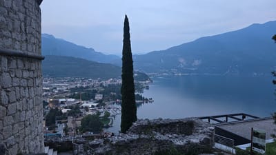 The image depicts a panoramic view of Riva del Garda, Italy, from a high vantage point. The scene is dominated by a large, dark green cypress tree standing tall and slender, bisecting the frame vertically. In the background, a calm, deep blue lake stretches out, reflecting the soft light of dusk. Across the lake, the town of Riva del Garda is visible, with its buildings clustered along the shoreline, illuminated by a scattering of lights. Mountains form a majestic backdrop, their slopes covered in dark green vegetation and their peaks fading into the pale blue sky. The overall atmosphere is serene and tranquil.

On the left side of the image, a stone wall and some greenery suggest the viewpoint is from an elevated structure, possibly a historical fortification or a scenic overlook. A signpost with some text is partially visible. The right side of the image is also bordered by a rough stone wall, consistent with a historical building.

The time of day appears to be twilight, with the sky transitioning from daylight to night. The weather seems clear and calm, contributing to the peaceful mood of the scene. The location cues, such as the architecture of the town and the surrounding mountainous landscape, strongly indicate Riva del Garda, Italy.