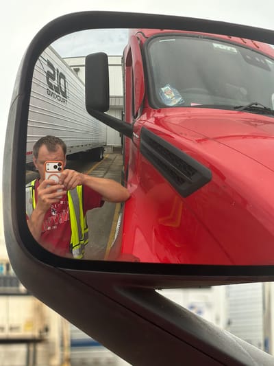 A person is taking a selfie in the side mirror of a red semi-truck. The person is wearing a red shirt and a high-visibility yellow vest. They are holding an orange-cased smartphone in their right hand. In the reflection of the mirror, other semi-trucks and trailers are visible, along with a building structure in the background. The ground appears to be wet, suggesting recent rain. The scene takes place outdoors in what appears to be a truck yard or industrial area. The image has a slightly overcast sky. The location is not explicitly stated in the image.