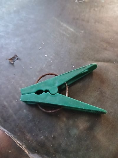 A single, green plastic clothespin with a rusty metal spring lies on a dark, textured surface. The clothespin is positioned diagonally across the frame, with its jaws open. A small, dried leaf or debris is visible to the left of the clothespin. The background is dark and appears to be an outdoor surface, possibly pavement or a tarp. No people or specific Hlushky, Ukraine landmarks are identifiable in this image.