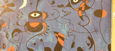 The image shows a close-up of a blue mural with abstract shapes and lines in orange, black, and white. The mural appears to be textured, with a slightly rough, speckled surface. There are several distinct elements: a large, stylized eye with a yellow iris and a white pupil within a purple and orange structure on the left side. Scattered throughout are star-like shapes with thin, dark lines radiating from a central dot. Black, curving lines intertwine with the other elements, creating a sense of movement and complexity. Orange shapes, some triangular or checkered, are interspersed with the black and blue. The overall style is reminiscent of surrealist or abstract art. A sliver of a yellow vertical surface with some warning labels is visible on the far right edge of the image.
This mural is located on the side of the "Palazzo dei Carmelitani" in Marsala, Italy, specifically on the wall facing Via Friscia.